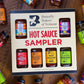 Butterfly Bakery of Vermont hot sauce sampler box with various hot sauce bottles around it on a wooden surface.