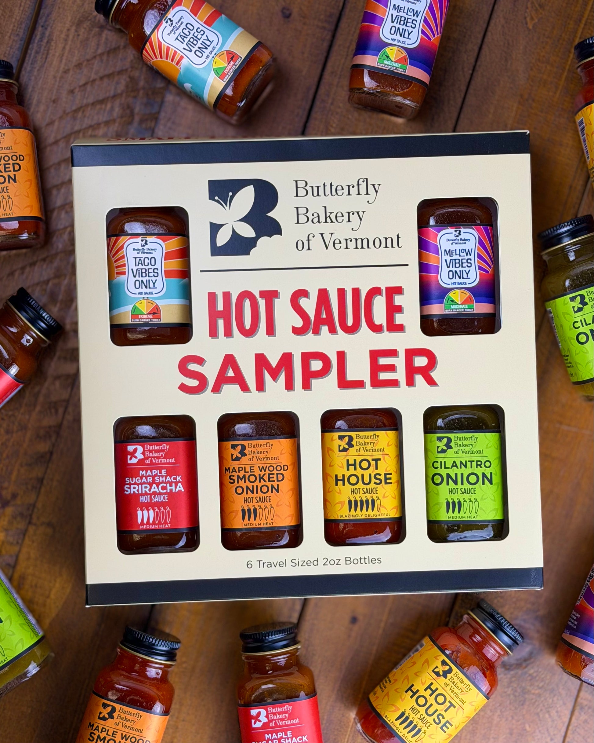 Butterfly Bakery of Vermont hot sauce sampler box with various hot sauce bottles around it on a wooden surface.