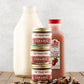 Three stacked jars of Fat Toad Farm caramel in front of a bottle of apple cider and a bottle of milk on a wooden surface with a light gray background.