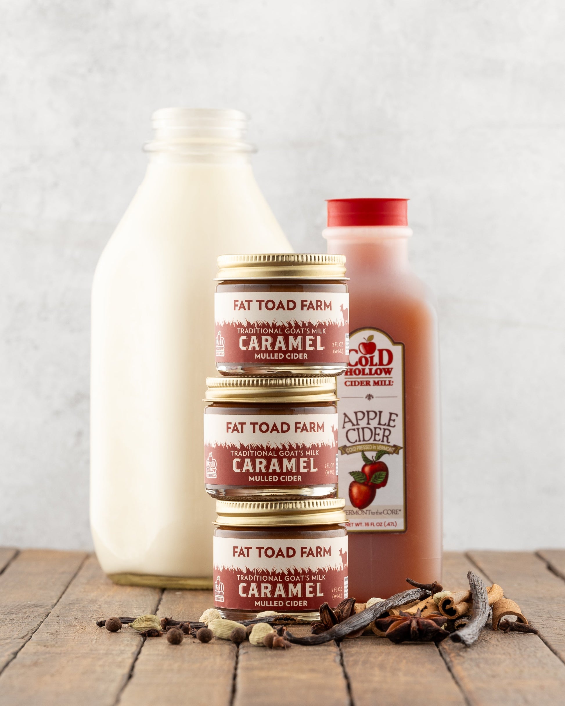 Three stacked jars of Fat Toad Farm caramel in front of a bottle of apple cider and a bottle of milk on a wooden surface with a light gray background.