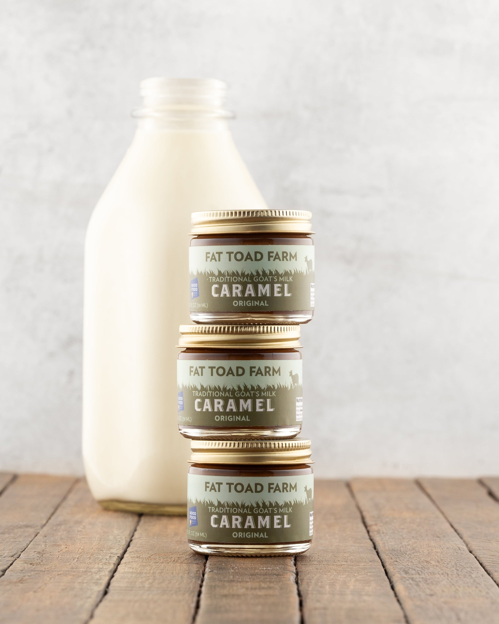Stacked 2oz jars of Fat Toad Farm Original Caramel Sauce in front of a bottle of milk on a wood surface