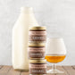Three jars of Fat Toad Farm Caramel stacked on a wooden surface with a bottle of milk and a glass of bourbon in the background.