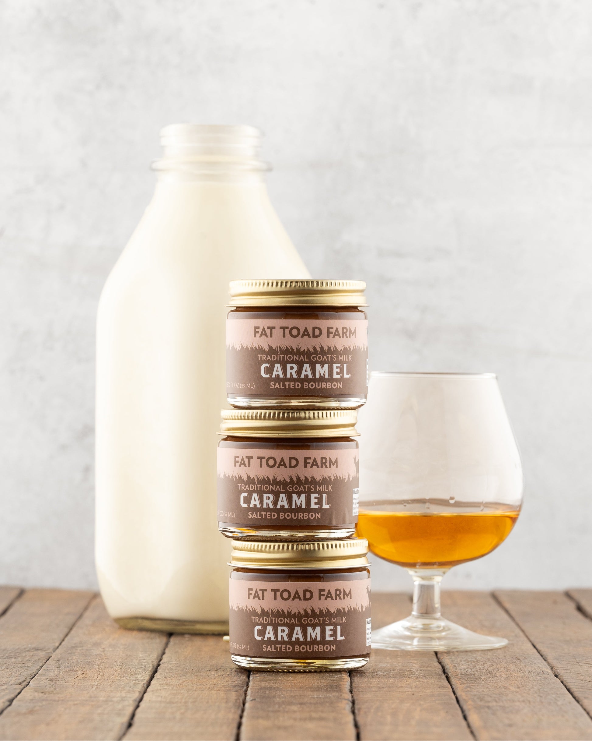 Three jars of Fat Toad Farm Caramel stacked on a wooden surface with a bottle of milk and a glass of bourbon in the background.