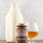 Jar of Fat Toad Farm Salted Bourbon caramel on a wooden table in front of a bottle of milk, and glass of bourbon