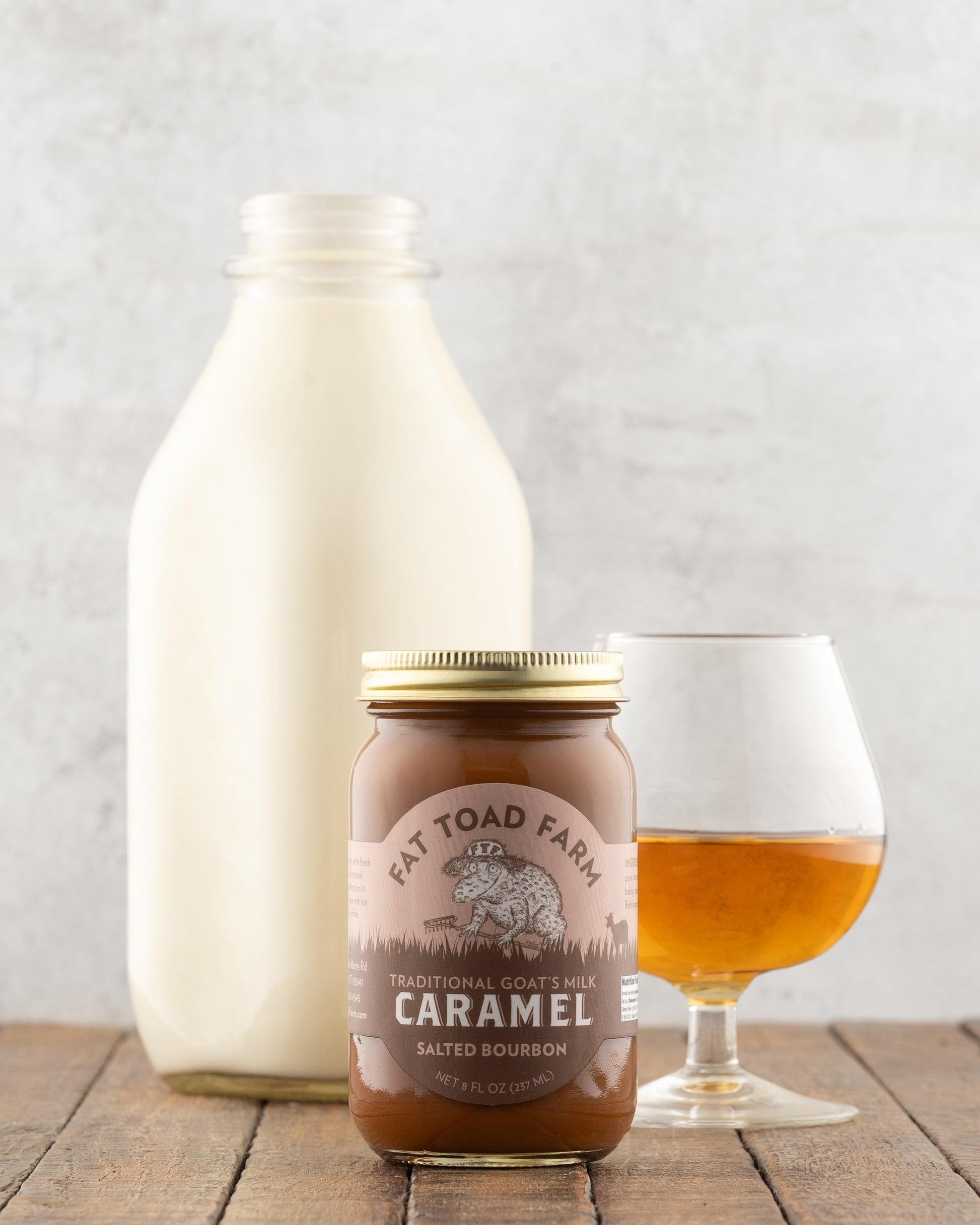 Jar of Fat Toad Farm Salted Bourbon caramel on a wooden table in front of a bottle of milk, and glass of bourbon