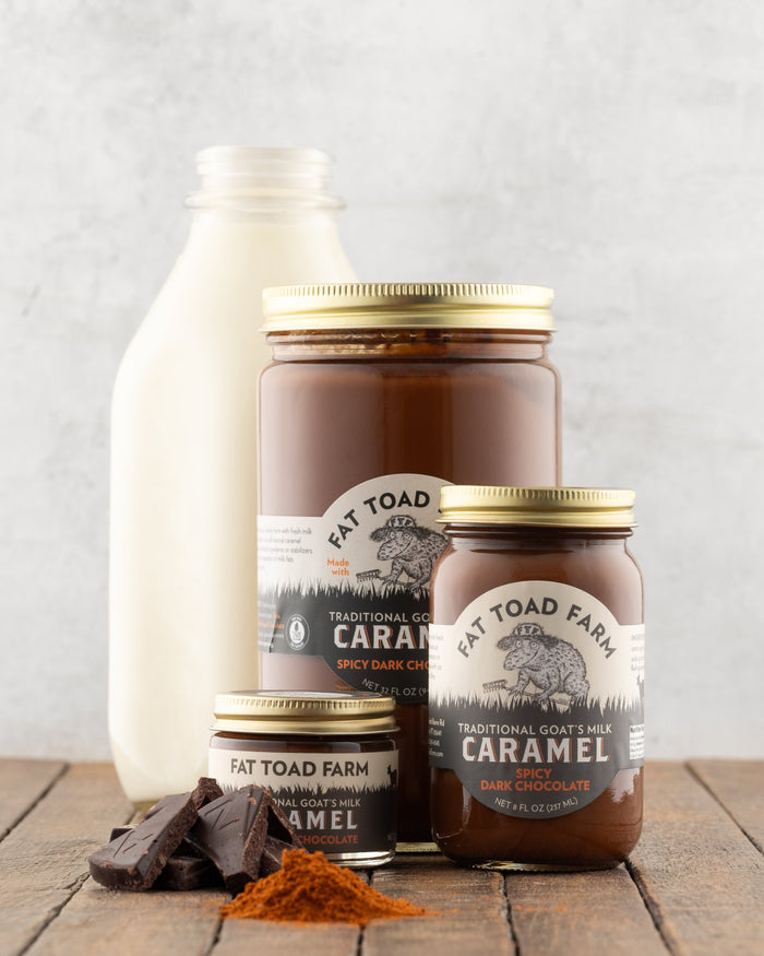 Fat Toad Farm Spicy Dark Chocolate caramel jars on a wooden surface with a bottle of milk, chunks of chocolate and ground cayenne pepper