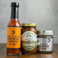 A jar of Heady Topper mustard, a jar of Fat Toad Farm Original caramel and a bottle of Maple Wood Smoked Onion hot sauce on a wooden surface with a gray background.