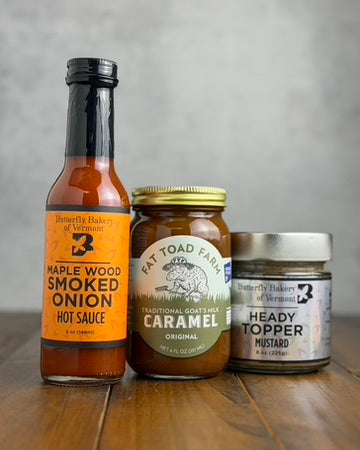 A jar of Heady Topper mustard, a jar of Fat Toad Farm Original caramel and a bottle of Maple Wood Smoked Onion hot sauce on a wooden surface with a gray background.