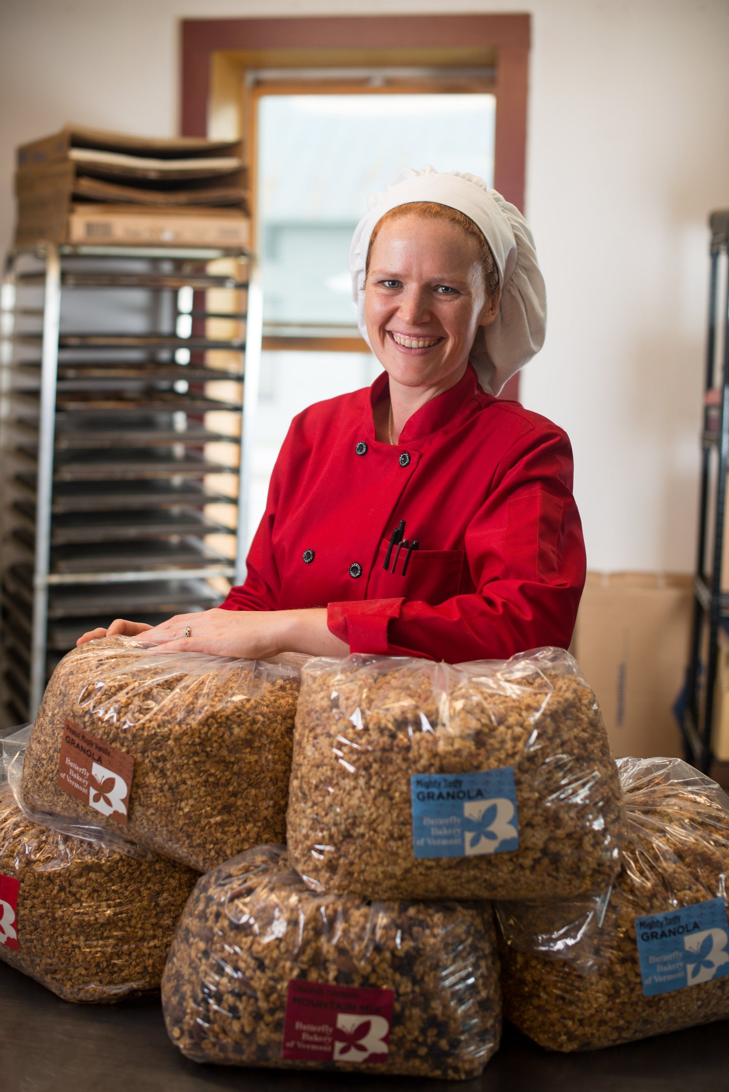 Claire Georges in a red chef's jacket standing behind bags of granola in a small commercial kitchen.