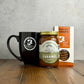 Black mug with a Butterfly Bakery of Vermont logo, a box of Maple Spice snaps, and a jar of Fat Toad Farm Original Caramel on a wooden surface.