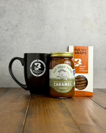 Black mug with a Butterfly Bakery of Vermont logo, a box of Maple Spice snaps, and a jar of Fat Toad Farm Original Caramel on a wooden surface.