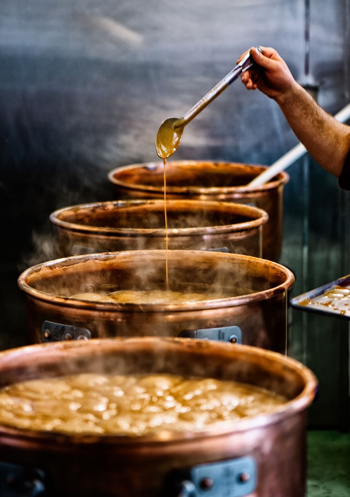 Person ladling caramel from a large copper pot into another, with a dark background.