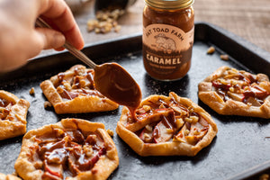 Person drizzling caramel sauce onto small pastries on a baking tray with a jar of Fat Toad Farm Caramel in the background.