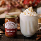 Jar of Fat Toad Farm caramel sauce next to a mug of hot chocolate with whipped cream on a wooden surface.