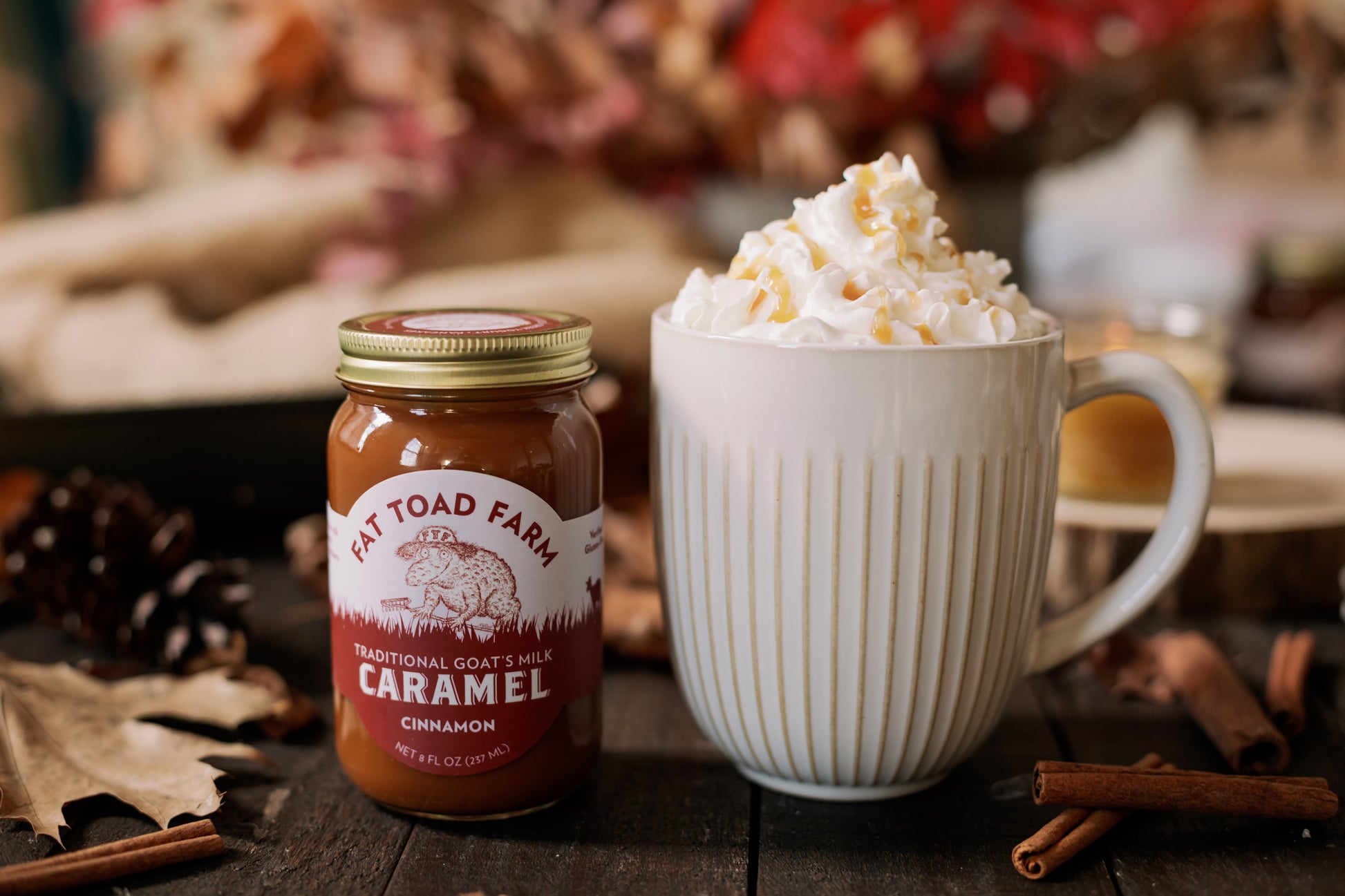 Jar of Fat Toad Farm caramel sauce next to a mug of hot chocolate with whipped cream on a wooden surface.
