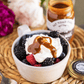 Bowl of dessert with berries and whipped cream, topped with a drizzle of caramel sauce, on a woven mat.