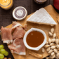 Platter with prosciutto, brie, grapes, nuts and a bowl of caramel sauce on a wooden board.