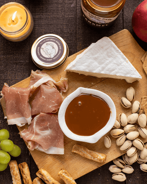 Platter with prosciutto, brie, grapes, nuts and a bowl of caramel sauce on a wooden board.