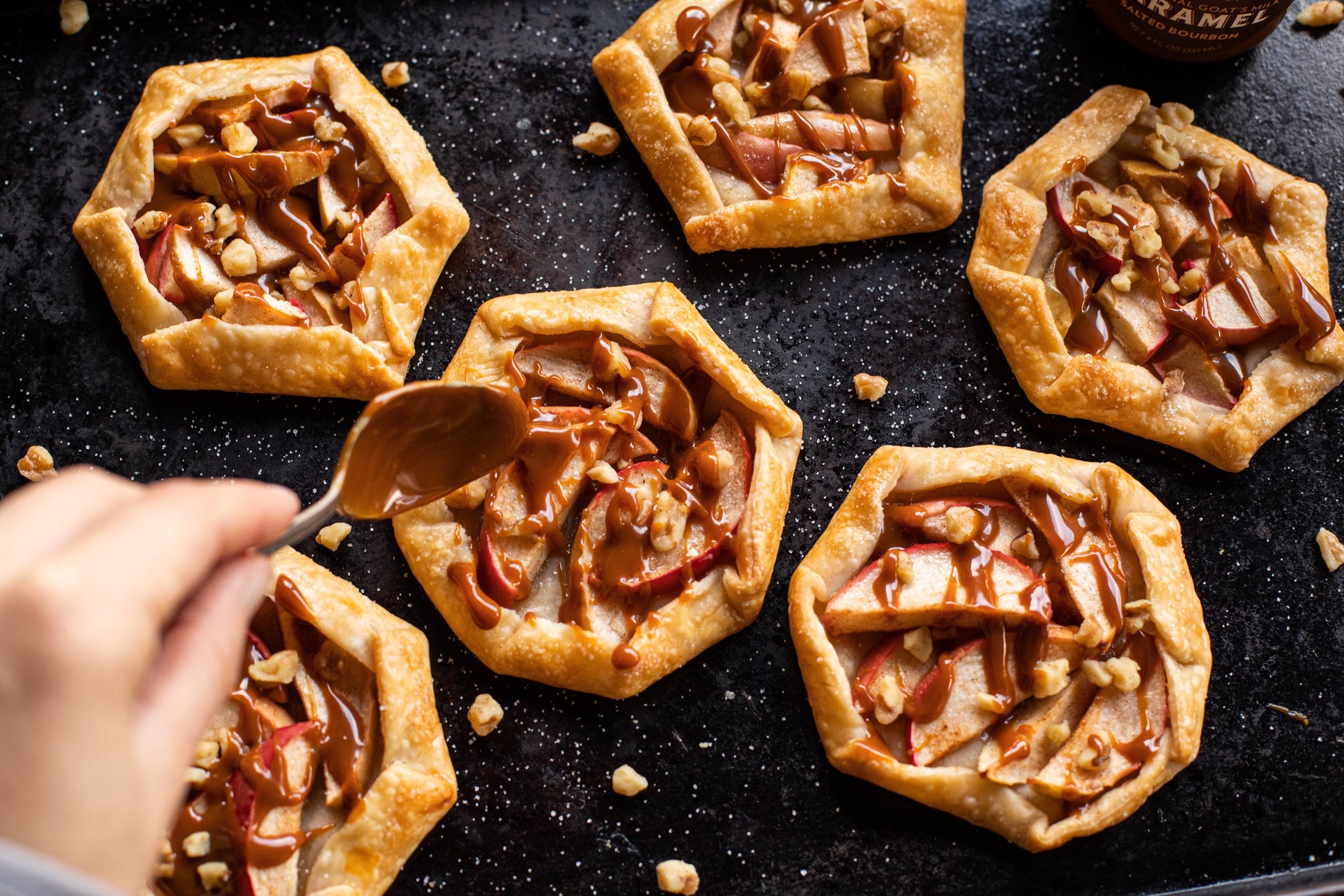 Small apple tarts with caramel sauce on a dark surface, being topped with a spoonful of caramel.