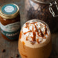 Glass of iced coffee with whipped cream, jar of caramel sauce, and jar of coffee beans on a wooden surface.