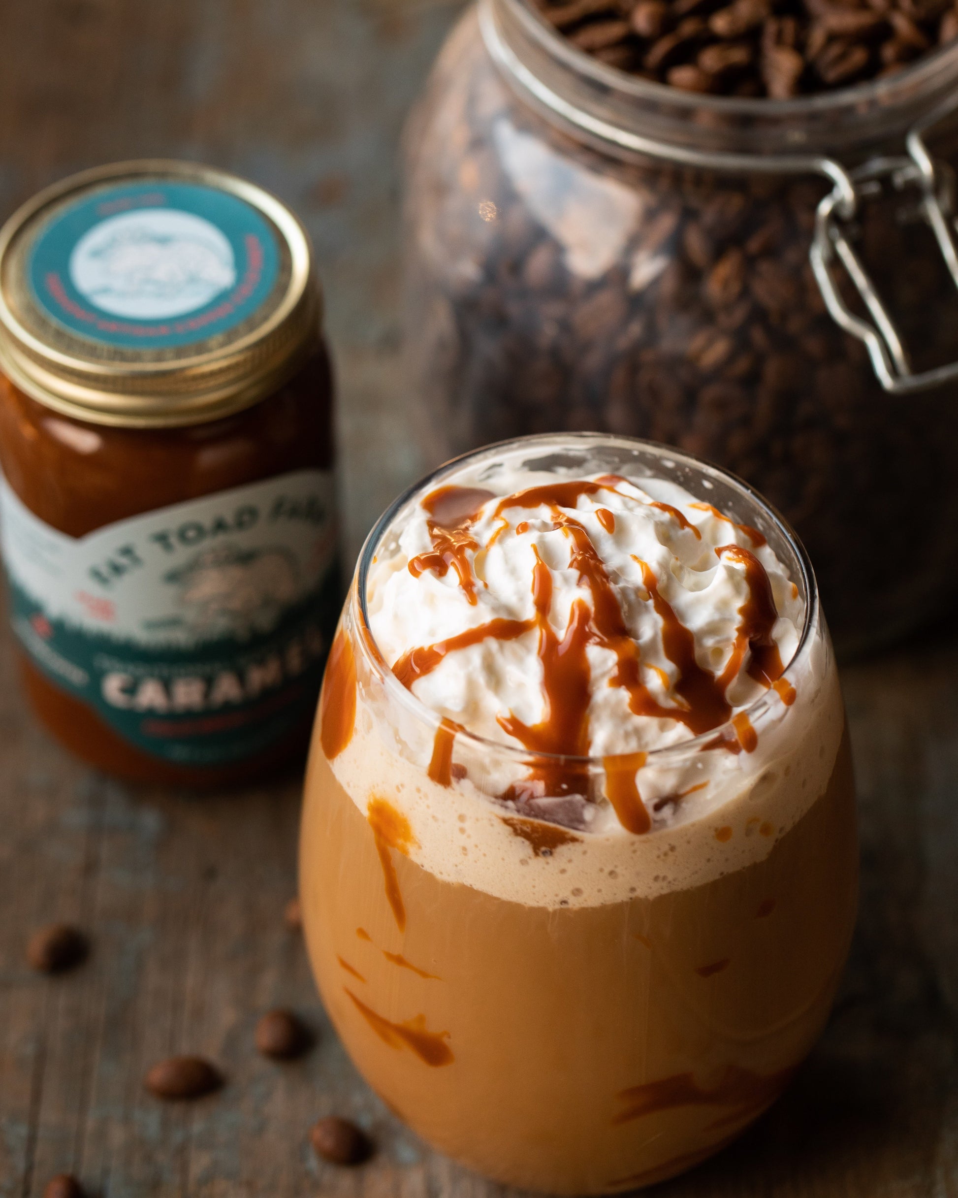 Glass of iced coffee with whipped cream, jar of caramel sauce, and jar of coffee beans on a wooden surface.