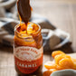 Jar of caramel from Fat Toad Farm with a spoonful being poured, surrounded by fruits on a wooden surface.