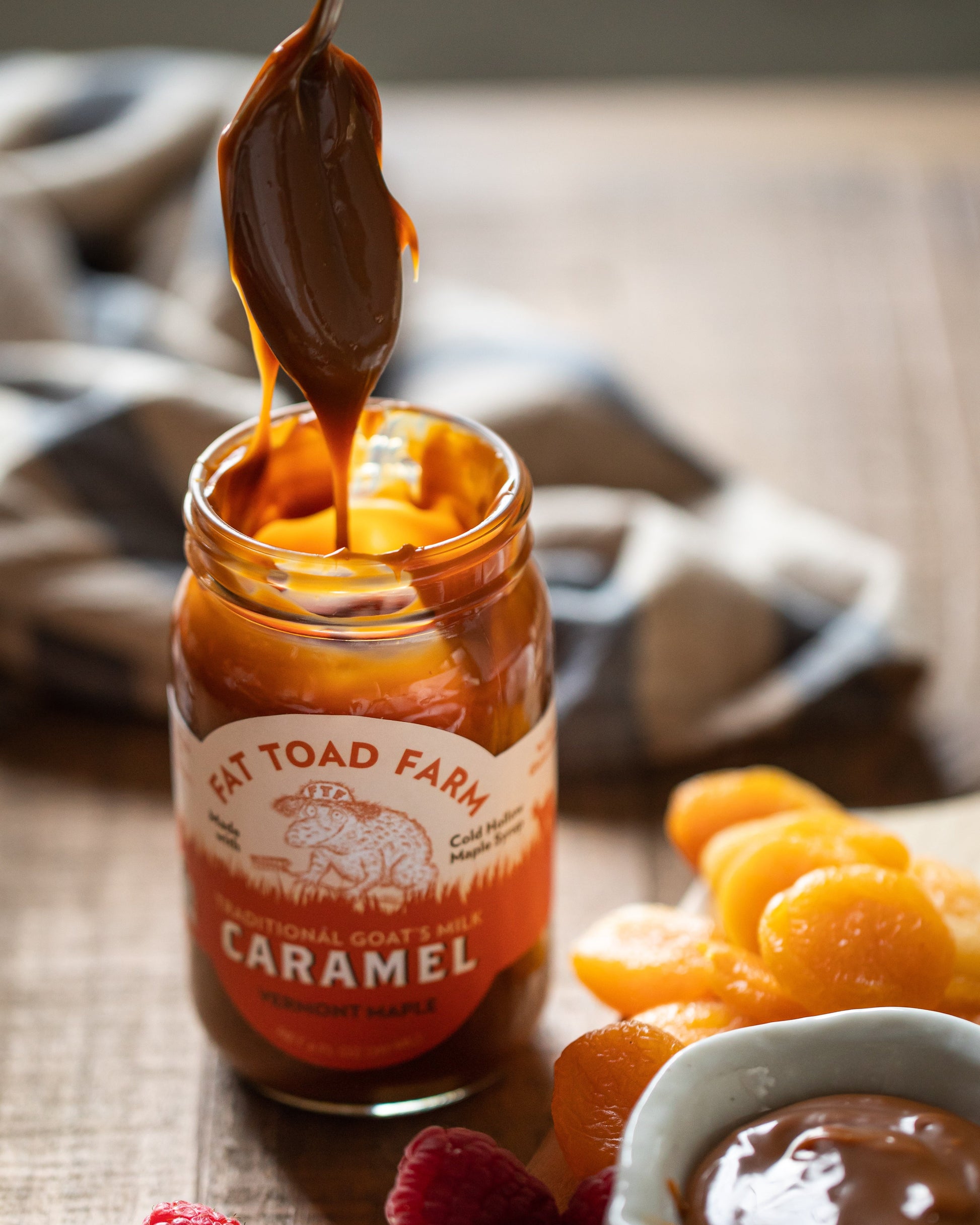 Jar of caramel from Fat Toad Farm with a spoonful being poured, surrounded by fruits on a wooden surface.