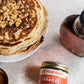 Stack of pancakes with nuts, a jar of Fat Toad Farm caramel sauce, and a bottle of maple syrup on a light surface.