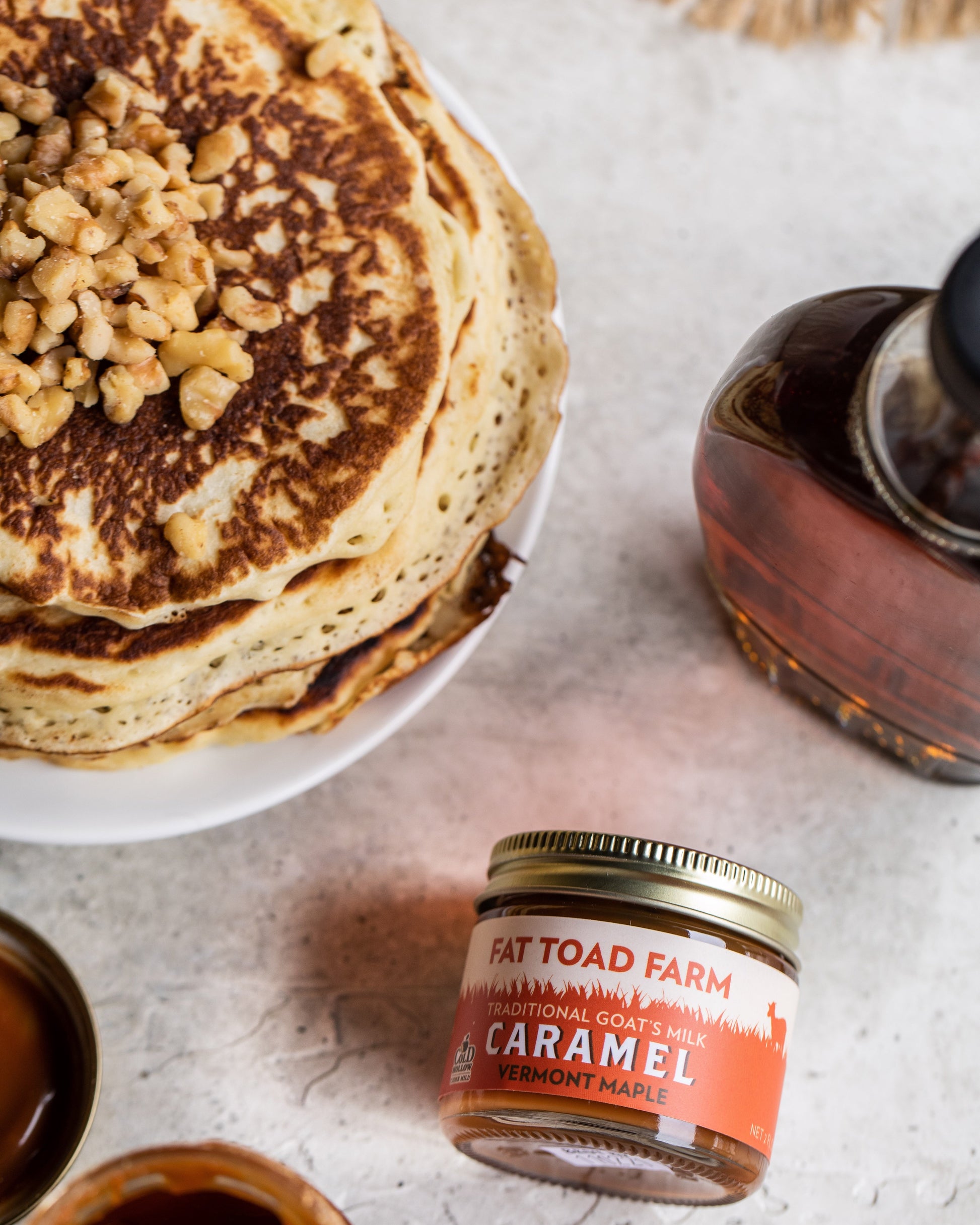 Stack of pancakes with nuts, a jar of Fat Toad Farm caramel sauce, and a bottle of maple syrup on a light surface.