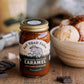 Jar of Fat Toad Farm caramel sauce with a bowl of ice cream and chocolate pieces on a wooden surface.