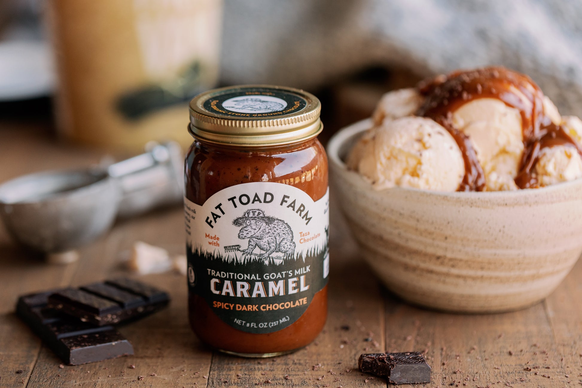 Jar of Fat Toad Farm caramel sauce with a bowl of ice cream and chocolate pieces on a wooden surface.