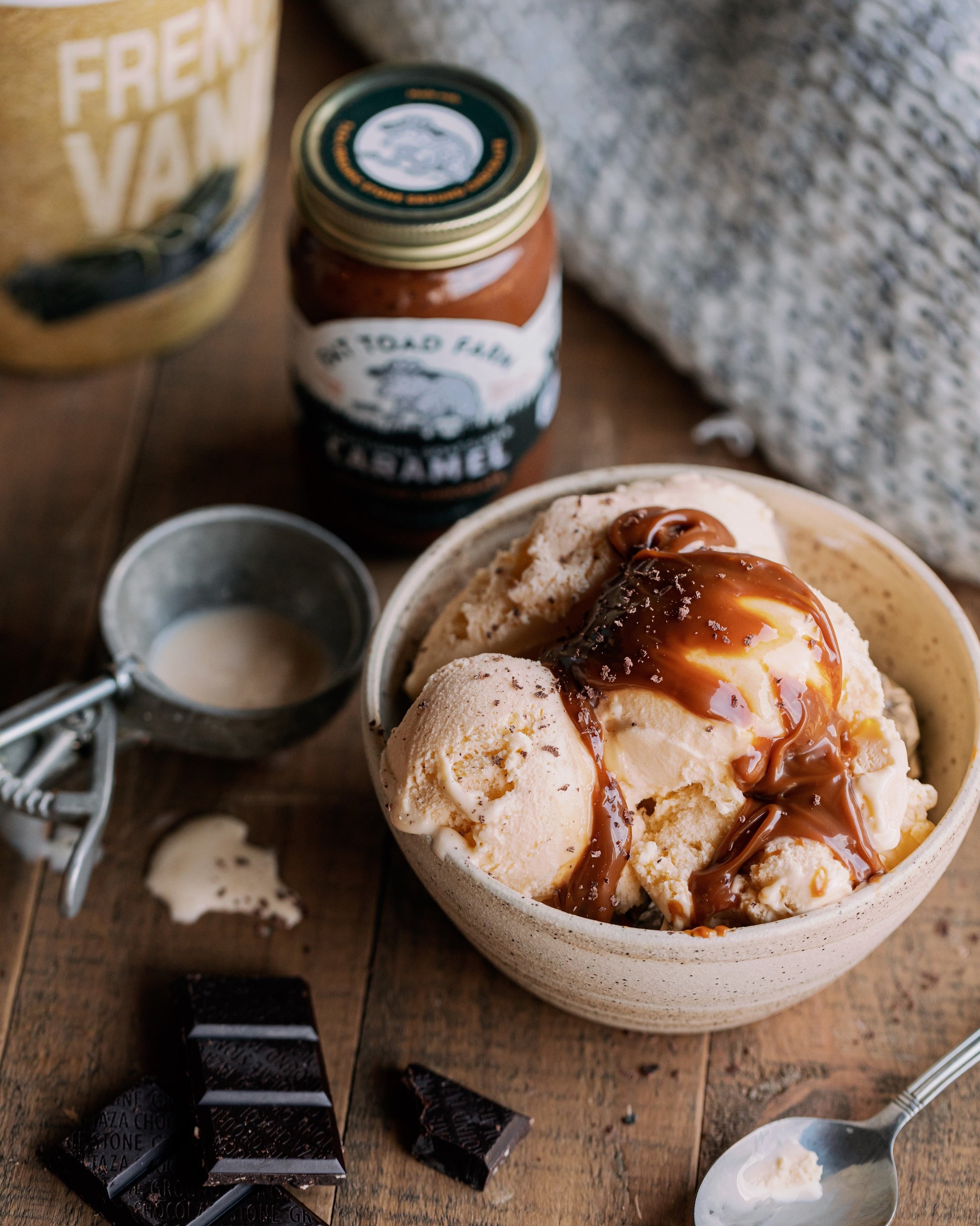 Bowl of ice cream with caramel sauce on a wooden surface with a jar of caramel sauce and a container labeled 'French Vanilla Ice Cream'.