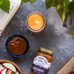 Candle, jar of caramel sauce, apples, and a book on a gray surface with green leaves.