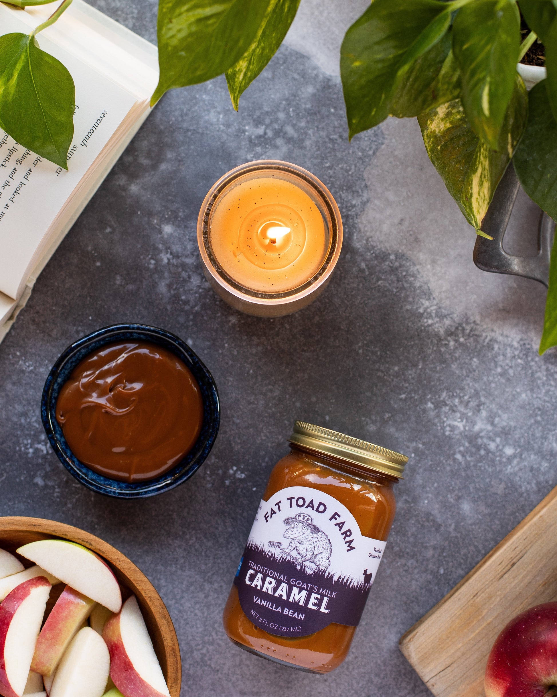 Candle, jar of caramel sauce, apples, and a book on a gray surface with green leaves.