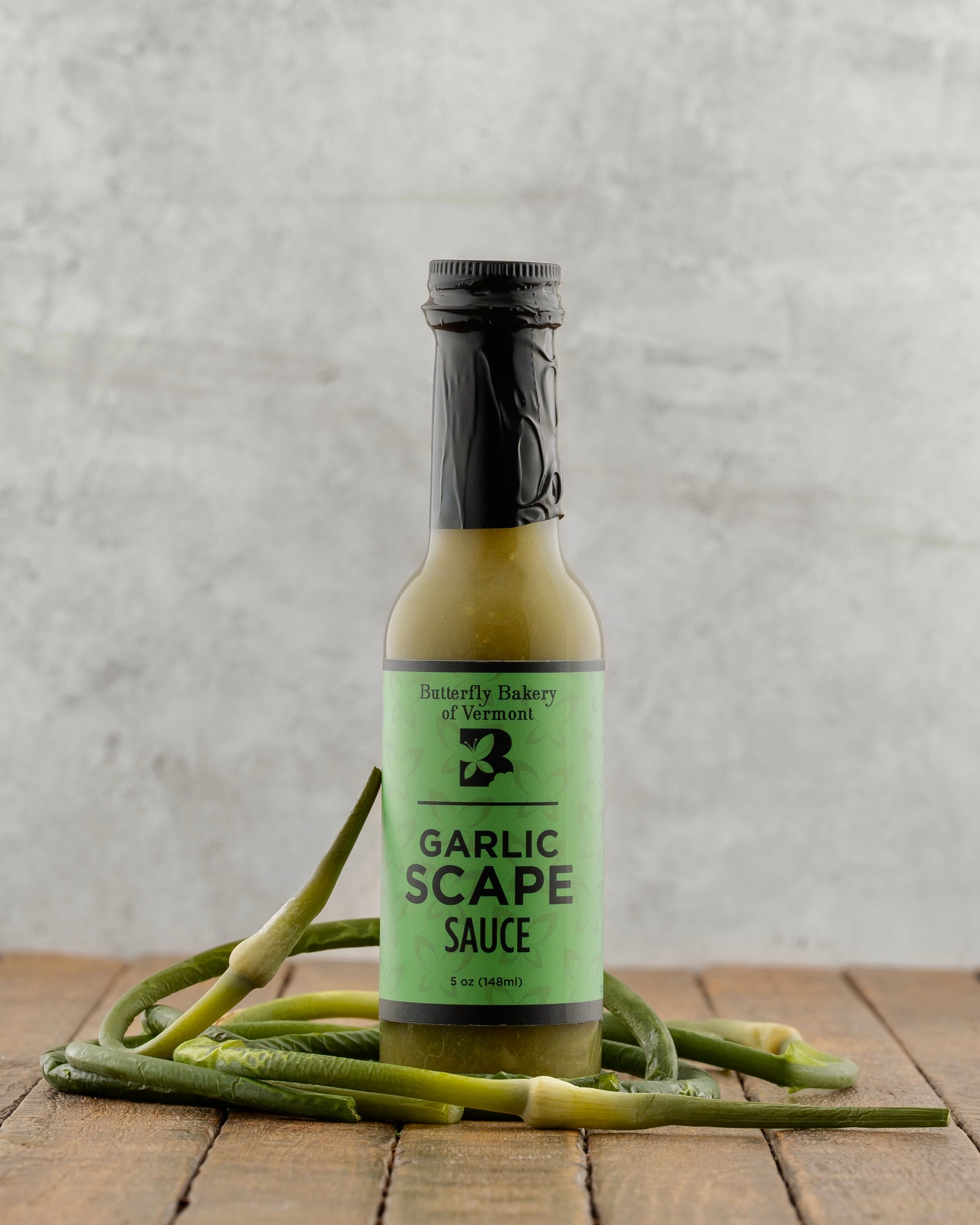 Bottle of Garlic Scape Sauce from Butterfly Bakery of Vermont on a wooden surface with garlic scapes.
