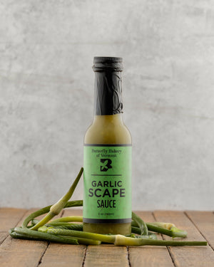 Bottle of Garlic Scape Sauce from Butterfly Bakery of Vermont on a wooden surface with garlic scapes.