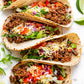 Tacos with ground beef, vegetables, cheese and hot sauce on a white surface with lime wedges.