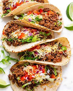 Tacos with ground beef, vegetables, cheese and hot sauce on a white surface with lime wedges.