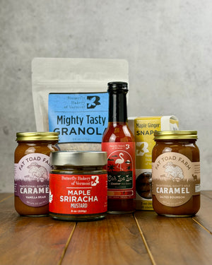 Two jars of Fat Toad Farm caramel, along with a bag of granola, a box of snaps, a bottle of hot sauce and a jar of mustard from Butterfly Bakery of Vermont on a wooden surface with a gray background