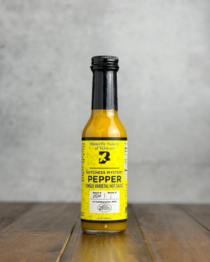 Bottle of Dutchess Mystery Pepper hot sauce on a wooden surface with a gray background