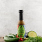 Bottle of Butterfly Bakery of Vermont Ghost Verde Pickle Hot Sauce on a wood surface next to fresh dill, garlic scapes, cucumber and hot peppers