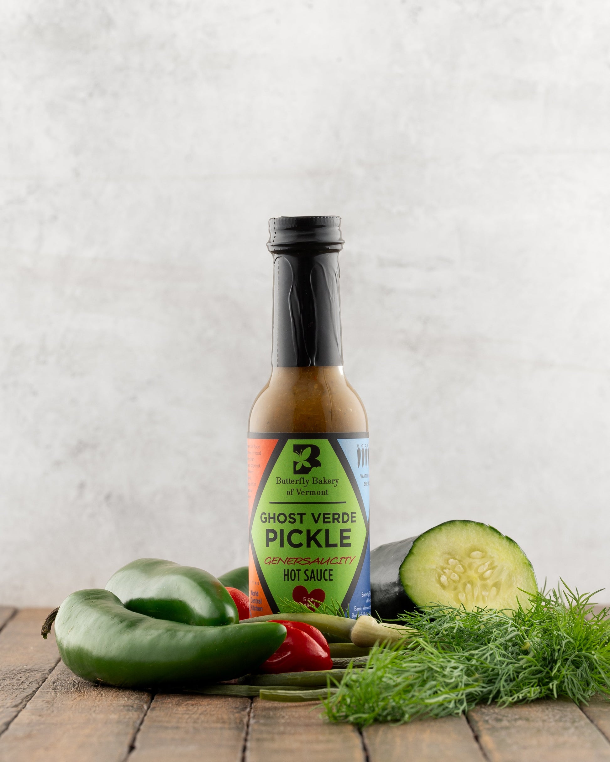 Bottle of Butterfly Bakery of Vermont Ghost Verde Pickle Hot Sauce on a wood surface next to fresh dill, garlic scapes, cucumber and hot peppers