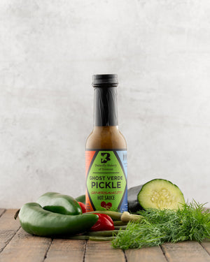 Bottle of Butterfly Bakery of Vermont Ghost Verde Pickle Hot Sauce on a wood surface next to fresh dill, garlic scapes, cucumber and hot peppers