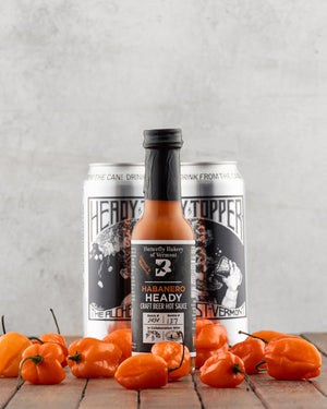 Butterfly Bakery of Vermont Habanero Heady Hot Sauce in front of two cans of Heady Topper beer on a wood surface and orange habanero peppers in the foreground