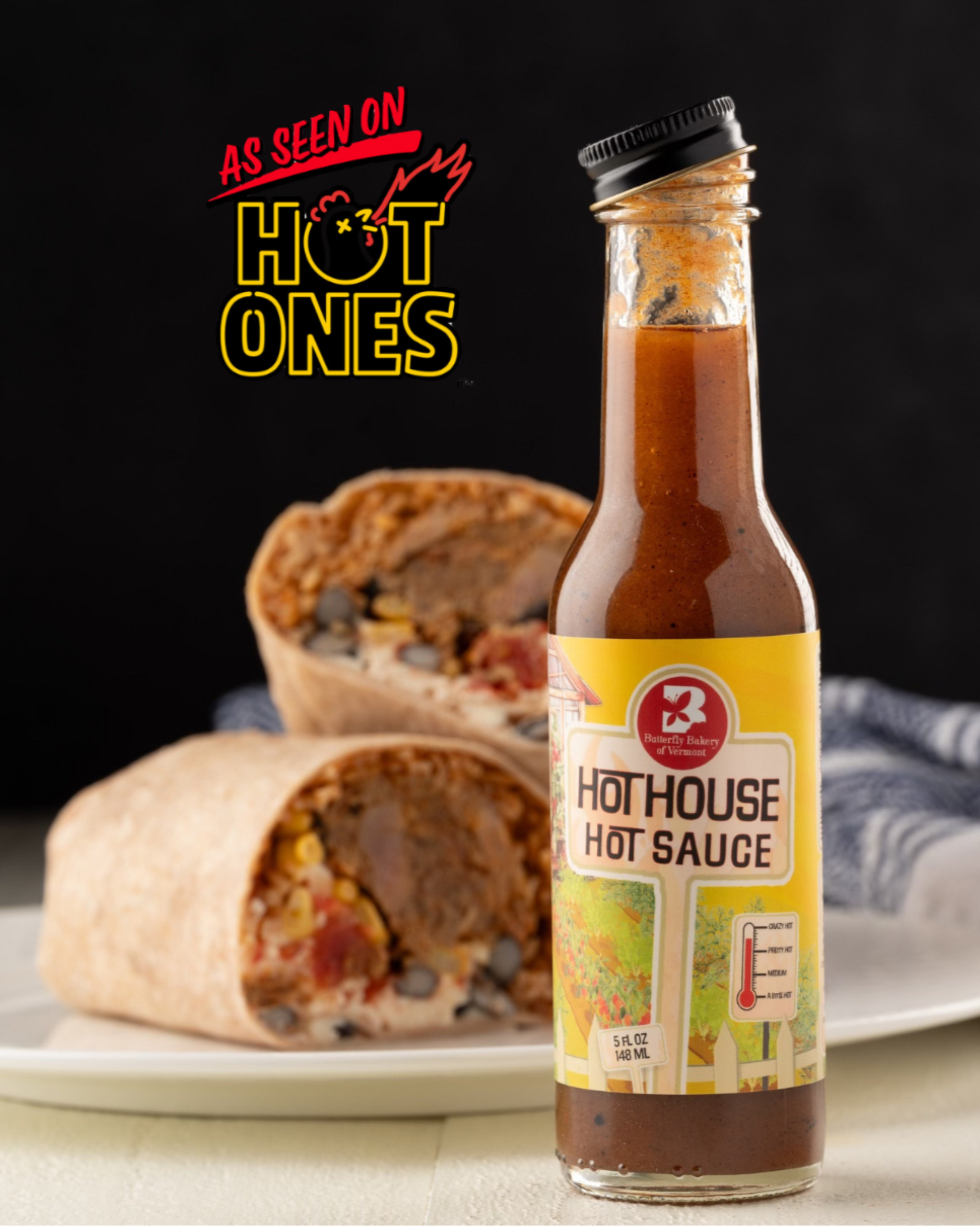 Bottle of Butterfly Bakery of Vermont Hot House hot sauce with a 'As Seen on Hot Ones' badge sitting on a table alongside a burrito cut in half on a plate