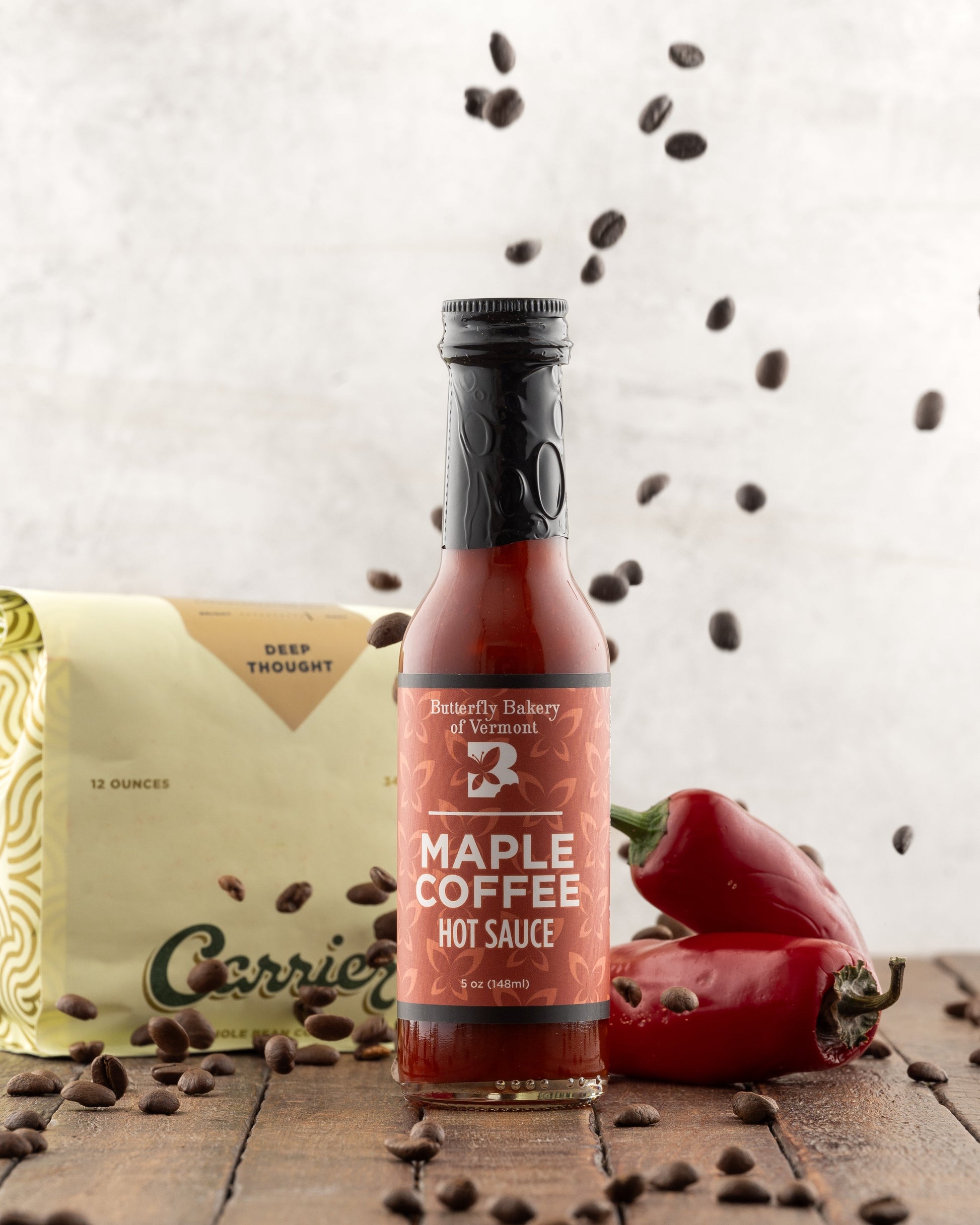 Bottle of Butterfly Bakery of Vermont Maple Coffee Hot Sauce on a wood surface and a bag of coffee in the background, with coffee beans being sprinked from above