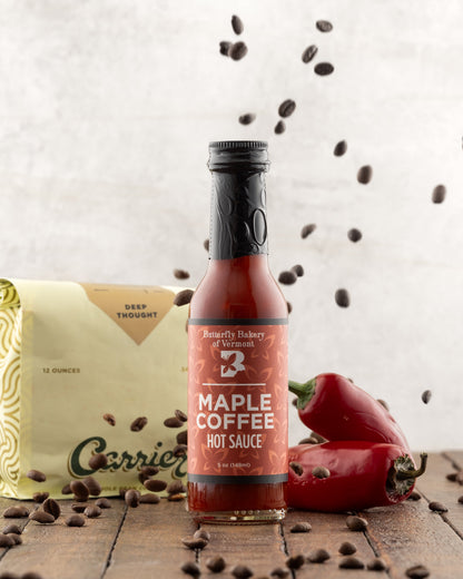 Bottle of Butterfly Bakery of Vermont Maple Coffee Hot Sauce on a wood surface and a bag of coffee in the background, with coffee beans being sprinked from above