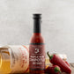 Bottle of Butterfly Bakery of Vermont Maple Rum Chipotle Hot Sauce next to jalapenos and a bottle of rum on a wood table surface