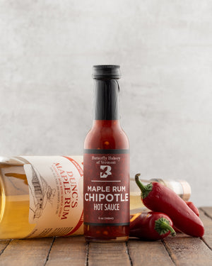 Bottle of Butterfly Bakery of Vermont Maple Rum Chipotle Hot Sauce next to jalapenos and a bottle of rum on a wood table surface
