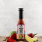 Bottle of Butterfly Bakery of Vermont Mellow Vibes Only Hot Sauce on a wood surface alongside red jalapenos, garlic scapes and lime wedges